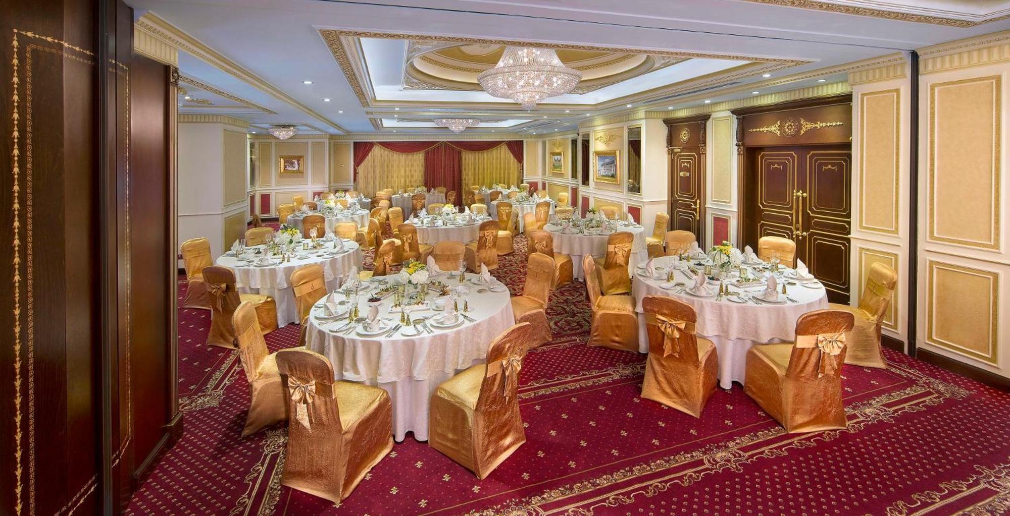 皇家玫瑰酒店 阿布扎比 外观 照片 The photo shows an elegantly arranged banquet hall set up for a formal event. There are several round tables covered with white tablecloths, each adorned with gold or cream-colored chair covers that match the upscale decor. The tables are set with fi